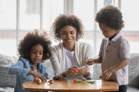 Happy African American young woman with and kids playing with colorful plasticine together, sitting on couch at home, smiling mum with adorable daughter and son involved in creative activityの写真素材