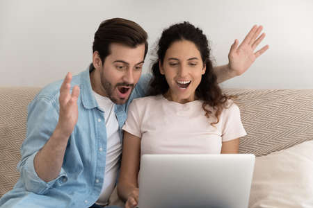 Amazed married couple splash hands receiving email message with profitable offer from online store. Excited surprised young spouses scream in delight look at laptop pc screen getting unexpected luckの写真素材