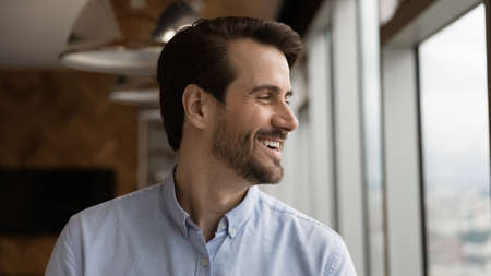 Head shot of positive optimistic man looking at large window with happy smile think of good new opportunities enjoy nice city view. Laughing young guy dreaming feeling confident of achieving successの写真素材