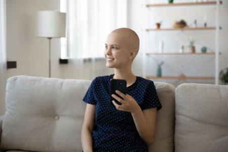 Happy millennial hairless woman struggle with oncology relax on sofa at home use modern smartphone gadget. Smiling young Caucasian female cancer patient survivor feel optimistic text on cellphone.の写真素材