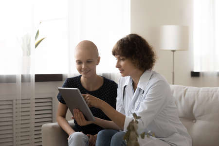 Caring doctor in white medical uniform and young Caucasian bald female cancer patient look at tablet screen discuss good results. Happy GP and smiling woman with oncology excited with recovery.の写真素材