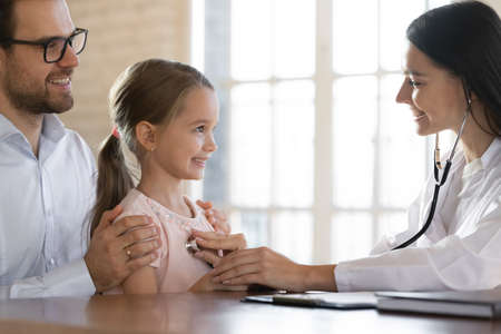 Close up of caring smiling female cardiologist listen to small girl child patient heart at hospital visit with dad. Woman doctor or nurse examine little kid at clinic consultation. Healthcare concept.の写真素材