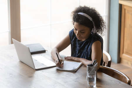 Top view African American woman in headphones taking notes, using laptop, listening to online...