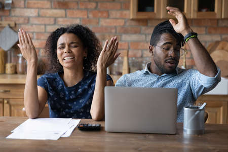 Close up unhappy African American couple frustrated by money problem, unexpected debt, bankruptcy, upset confused wife and husband sitting at table with laptop, calculating bills, household expensesの写真素材