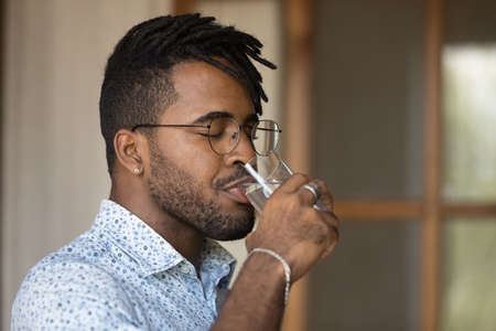 Close up side view thirsty African American man wearing glasses enjoying fresh pure mineral water with closed eyes, holding glass, healthy lifestyle and good daily habit concept, body and skin careの写真素材