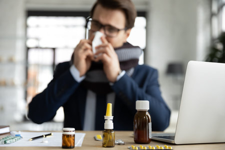 Number of medicines or pills bottles on office desk, sick male employee work in background. Close up...