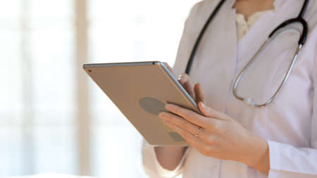 Close up young female doctor holding digital computer tablet in hands, web surfing information or consulting patient online, providing medical service, copy space, modern tech and medicine concept.の写真素材