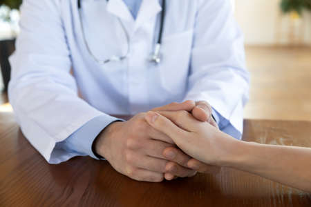 Close up kind male gp doctor holding hand of female patient showing empathy, supporting at clinic...