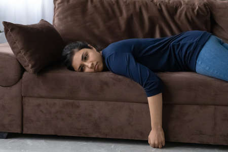 Tired young Indian woman lying on couch in living room suffer from depression or anxiety. Exhausted...