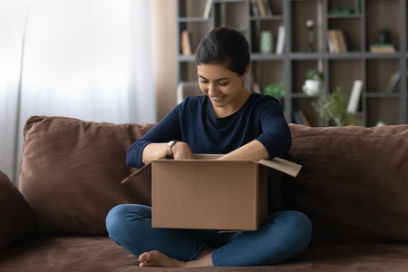 Happy millennial Indian woman relax at home unbox cardboard package with internet order at home. Smiling young ethnic female buyer unpack box shopping online. Good delivery service, shipping concept.の写真素材