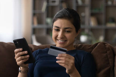 Happy millennial Indian woman relax at home shopping online on smartphone paying with credit card....