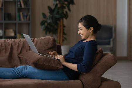 Happy millennial Indian woman rest on sofa at home look at laptop screen study distant on gadget. Smiling young mixed race female work online on computer, relax on couch in living room with device.の写真素材