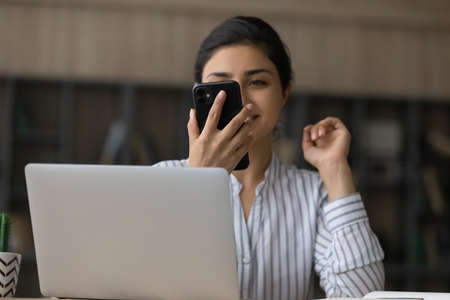 Happy millennial Indian woman work on computer use modern cellphone gadget talk on video call....