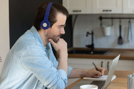 Pensive Caucasian male in earphones look at laptop screen writing studying online on laptop from...
