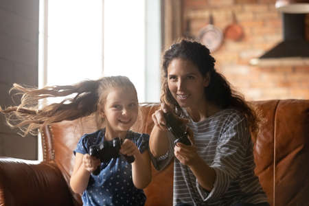 Happy young mother and little daughter playing video game, holding remote controllers, family...