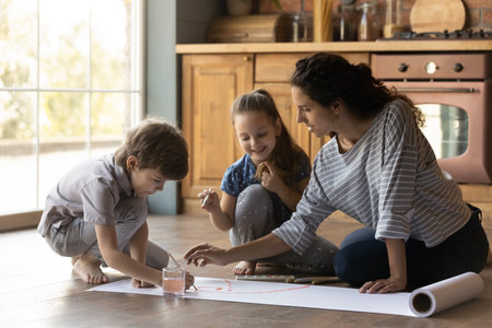 Caring mother and little kids painting colorful watercolors at home together, sitting on warm wooden...