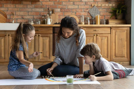 Caring young mother and two kids playing, painting colorful watercolor, sitting on warm wooden floor...