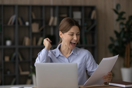 Overjoyed young Caucasian woman work on computer at home office triumph read good news in postal paperwork. Excited businesswoman feel euphoric with promotion get post letter correspondence.の写真素材