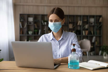 Young Caucasian businesswoman wear facemask work online on computer at home office during covid-19...
