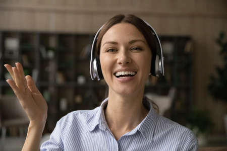 Close up portrait of overjoyed millennial Caucasian woman in earphones greeting talking on video...