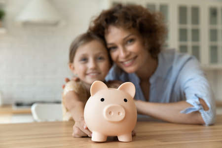 Take care of future. Blurred portrait of smiling young mother small girl daughter look at camera show funny pig for coins offer you to make bank deposit save money. Focus on piggybank on kitchen tableの写真素材