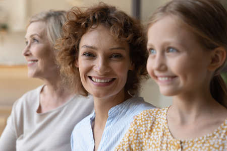 Generational similarity. Three generation family females relatives of diverse age old grandma adult mom daughter kid sit in row together. Focus on smiling face of young woman at centre look at cameraの写真素材