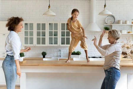 Active family of 3 female generations retired grandma adult mom little daughter grandchild have fun dance at modern kitchen. Energetic dynasty granny mom kid girl jump by music on funny party at homeの写真素材