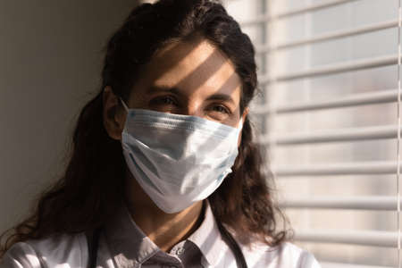 Head shot smiling young female doctor wearing protective medical face mask and uniform standing near window in hospital office, professional therapist physician looking to aside, disease preventionの写真素材