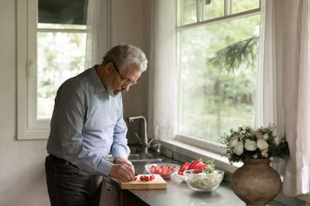 Mature adult man cooking dinner at home, cutting fresh vegetables for salad in kitchen. Old senior pensioner keeping healthy diet and lifestyle, taking care about nutrition, eating raw organic foodの写真素材