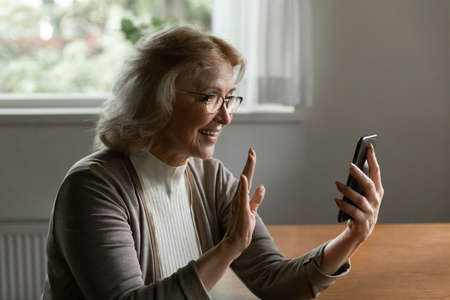 Happy middle aged pensioner woman with smartphone making video call, using mobile phone, waving...