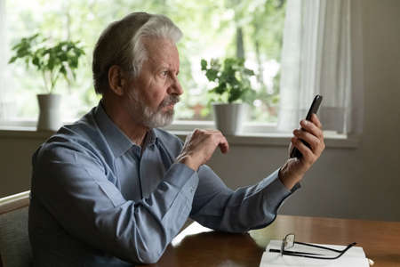 Serious mature 60 - 70s aged smartphone user reading message on screen. Senior man using app on mobile phone, making video call from home, receiving concerning news, checking email or social mediaの写真素材