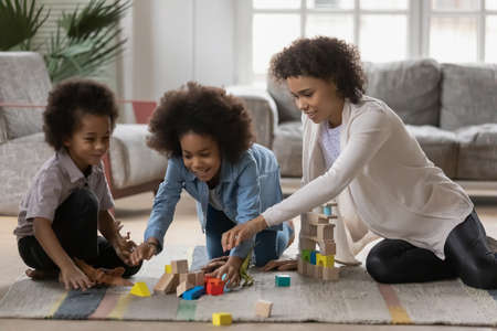 Happy black kids and mom building with wooden construction blocks playing on floor. African American babysitter watching children at home, teaching learning game for preschool. Daycare conceptの写真素材