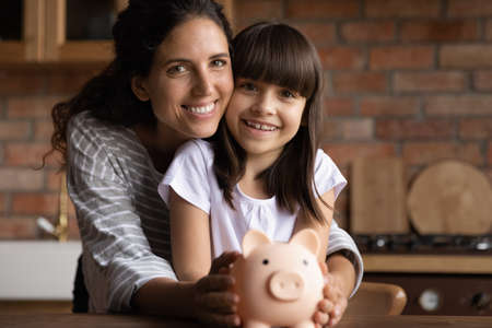 Portrait of happy Latino young mom and small 8s daughter recommend smart living and saving. Smiling Hispanic mother and little teen girl child hold piggybank feel economical making investment.の写真素材