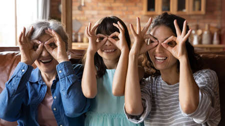 Portrait of overjoyed three generations of Latino women make glasses with fingers relax together at home. Happy young woman with small girl child daughter and mature grandmother have fun on weekend.の写真素材