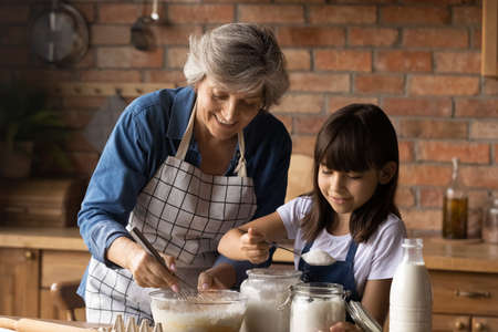 Smiling mature Hispanic granny and small granddaughter work with flour bake cookies in kitchen at home. Happy caring senior grandmother and little grandchild cook breakfast or dessert together.の写真素材