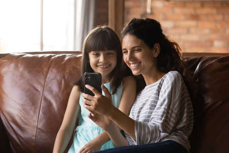 Smiling small Hispanic girl child and young mother look at cellphone screen speak on video call. Happy Latino mother and little daughter have webcam digital talk on smartphone. Virtual event concept.の写真素材