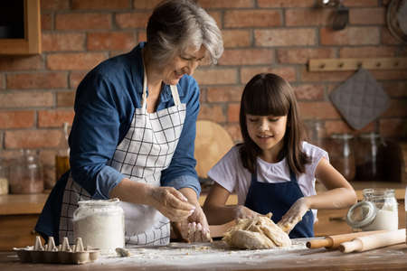 Happy little teen Latino girl child have fun baking making dough for cookies with elderly...