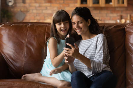 Happy little girl child relax on sofa at home with young Latino mother talk on video call on...