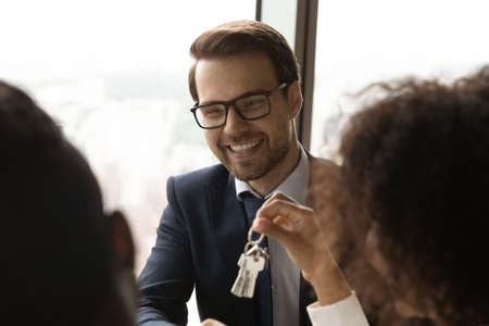 Close up smiling Caucasian realtor manager salesman wearing glasses giving keys to African American...