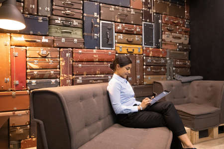 Confident Indian young businesswoman using tablet, sitting on couch in modern office, browsing apps, checking business email, working on online project, chatting with partners in social networkの写真素材