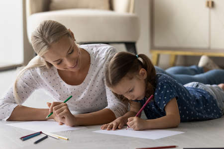 Loving happy young Caucasian mom and little daughter lying on floor drawing in album together. Caring smiling mother and small girl child have fun relax paining, involved in artistic activity.の写真素材