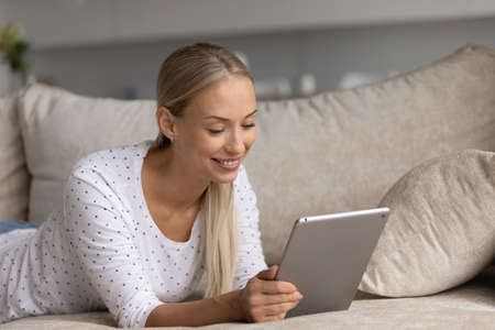 Smiling millennial Caucasian woman relax on couch at home browse internet on modern tablet. Happy...