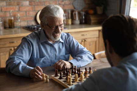 Smiling elderly Caucasian 60s father have fun play chess at home on weekend together with adult son. Happy old dad engaged in logical board game with grownup kid. Hobby, entertainment concept.の写真素材