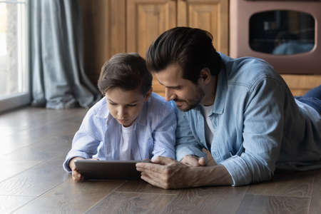Loving young Caucasian father and small preschooler son lying on floor at home have fun using modern tablet gadget together. Caring dad and little boy child enjoy browsing internet on pad device.の写真素材