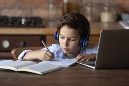 Smart little Caucasian boy in headphones sit at desk at home study online on computer write in notebook. Focused small kid child in earphones have webcam distant lesson on laptop. Education concept.の写真素材