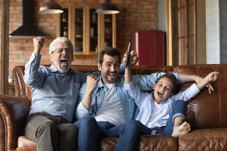 Overjoyed three generations of Caucasian men relax at home watching football celebrate win together. Happy boy with young father and old grandfather have fun cheering enjoy game match online.の写真素材
