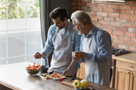 Happy elderly Caucasian father and adult son cook tasty dish together in home kitchen. Smiling older 60s dad and grownup man child prepare healthy vegetable salad for dinner. Diet, hobby concept.の写真素材