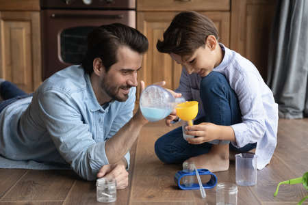 Caring young Caucasian father and little 7s son have fun do chemistry experiments at home in kitchen. Loving playful dad and small boy child mix ingredients play lab game together. Hobby concept.の写真素材