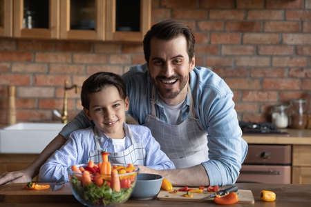Portrait of smiling young Caucasian father with small son have fun cooking tasty healthy vegetable salad for dinner. Happy dad and little boy child prepare delicious food at home kitchen together.の写真素材