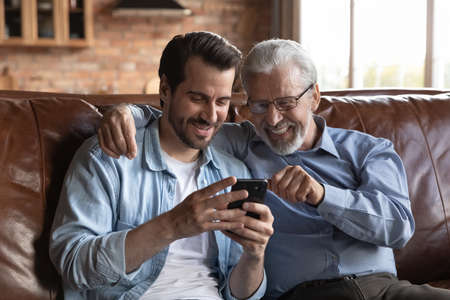 Smiling mature grey-haired 60s father and grownup son relax on sofa at home browse smartphone together. Happy older Caucasian dad and adult man child have fun use cellphone talk on call online.の写真素材
